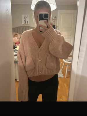Beige stickad cardigan med pärlknappar - Mysig beige stickad cardigan med v-ringning, croppad passform och stora fickor framtill. Tröjan har pärlknappar och långa ärmar med breda muddar. Perfekt för lager-på-lager och chill dagar.