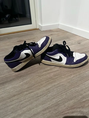 Nike Jordan 1 Low svart/lila/vit - Nike Jordan 1 Low sneakers i svart, lila och vitt med klassisk svart swoosh på sidan. Skorna har snörning, perforerad tå och platt sula. Perfekt för dig som gillar streetwear och vill sticka ut med en ikonisk modell.