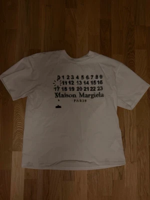 Vit Maison Margiela t-shirt - Vit t-shirt från Maison Margiela med det ikoniska numrerade trycket framtill. Klassisk passform och rund hals. Tillverkad i mjuk bomull för en skön känsla. Perfekt för dig som gillar minimalistisk och stilren design.