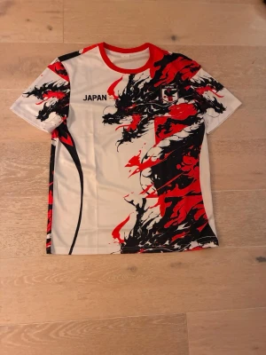 Japan sporttröja med drakmotiv - Säljer en vit sporttröja med röd och svart drakdesign, officiell Japan-logga på bröstet och texten 'JAPAN'. Tröjan har rund hals med röd kant och är tillverkad i ett lätt, snabbtorkande material som passar perfekt till träning eller match.