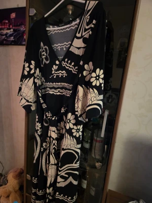 Svart och vit blommig långklänning - Unik långklänning från KappAhl i svart och vitt med blommigt och grafiskt mönster. Klänningen har v-ringning, vida trekvartsärmar och ett knytband i midjan för snygg passform. Perfekt för dig som gillar statement-plagg med retrovibbar.