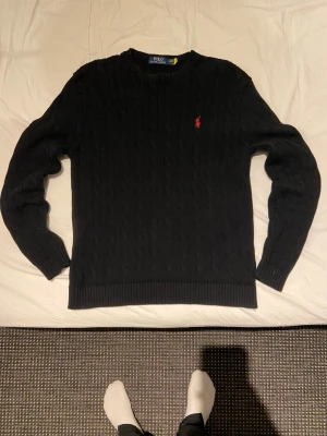 Svart kabelstickad tröja Polo Ralph Lauren - Kabelstickad svart tröja från Polo Ralph Lauren med rund halsringning och ribbade muddar. Liten röd broderad logotyp på bröstet. Klassisk och stilren design som passar perfekt till jeans eller chinos.pris går och  diskuteras ,destro snabbare affär destro billigare 