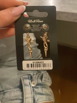 Guldiga örhängen med hjärta och pil - Säljer ett par guldiga örhängen från ur&penn med hjärtformad detalj och pil. Hjärtat har en ljusrosa sten. Örhängena är helt nya köpte de idag