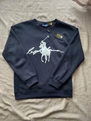 Mörkblå sweatshirt från Polo Ralph Lauren - Snygg mörkblå sweatshirt från Polo Ralph Lauren med stort vitt tryck av den klassiska polospelaren och loggan på bröstet. Tröjan har rund halsringning, ribbade muddar och är gjord i mjuk bomullsmix. Perfekt för en avslappnad och stilren look.