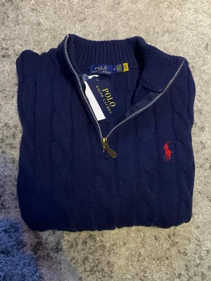 Marinblå Ralph Lauren quarter-zip - En elegant kabelstickad quarter-zip i blått med röd logga. Levererar behaglig upplevelse på, och en stilren look samtidigt. Storleken är M fast passar som en mindre S enligt mig, skicket är mycket bra eftersom den är näst intill oanvänd. Hör gärna av dig vid funderingar så löser vi det! 😁
