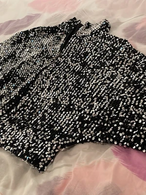 Svart paljett korsett med dragkedja - Säljer en svart korsett topp täckt med glittriga paljetter och dragkedja framtill. Perfekt för dig som vill sticka ut och glänsa lite extra. Sitter tight och ger en snygg siluett. Matcha med jeans eller kjol för en trendig look.