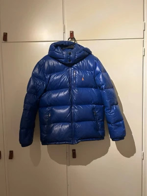 Blå dunjacka från Polo Ralph Lauren - Snygg blå dunjacka från Polo Ralph Lauren med huva och dragkedja framtill. Jackan har två sidofickor med dragkedja och den klassiska lilla loggan på bröstet. Perfekt för kalla dagar och riktigt skön att bära. Finns några fläckar. Se bild, annars övrigt bra skick. Storlek xl (junior) passar även som xs vuxen. 