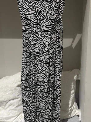 Zebramönstrad omlottklänning H&M storlek 38 - Snygg lång zebramönstrad omlottklänning från H&M i svart och vitt. Klänningen har v-ringning, knyt i midjan och är ärmlös. Perfekt för dig som vill sticka ut med ett djurmönster och en skön passform. Smala justerbara axelband