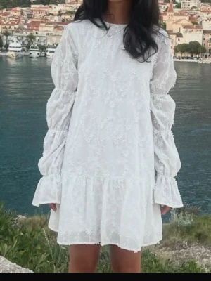 Vit blommig studentklänning  - Söt vit kortklänning med blommigt broderi och volangkant. Klänningen har långa puffärmar och är tillverkad i ett lätt tyg som ger en romantisk känsla. Perfekt för sommarens alla tillfällen och student! Den är aldrig använd och prislappen kvar!! Pris går att diskutera! 