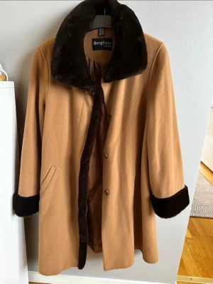 Camel färgad kappa från Berghaus med mörkbruna fuskpälsdetaljer - Berghaus fashion kappa i mycket fint skick. Den är gjord av ull och Kashmir. Storlek ca 38-40. Från rök och djurfritt hem.