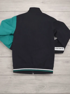Lacoste vindjacka i svart och turkos - Snygg vindjacka från Lacoste i svart med turkosa detaljer och vit rand över bröstet. Jackan har dragkedja, ribbade muddar med gröna och vita ränder samt två fickor framtill. Klassisk Lacoste-logga på bröstet och märkesdetalj på ärmen.