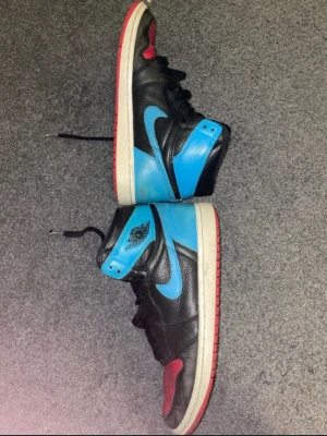 Nike Air Jordan 1 svart/blå/röd - Nike Air Jordan 1 sneakers i svart skinn med blå detaljer och röd tå samt yttersula. Klassisk hög modell med snörning och ikonisk vinge-logga på sidan. Perfekta för dig som vill sticka ut med färgkombon och streetwear-vibe.