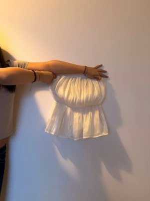 Vit axelbandslös tubtopp med volang - Säljer en söt vit axelbandslös tubtopp med volang och lätt glansigt tyg. Toppen har en bandeauform och är perfekt för sommardagar. Den är luftig och har en snygg rynkad effekt upptill.
