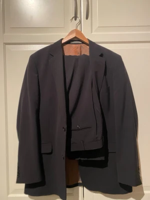 Mörk kostym från Oscar Jacobson - Snygg mörkgrå kostym från Oscar Jacobson med diskret randigt mönster. Klassisk kavaj med två knappar och matchande byxor. Perfekt för dig som vill ha en stilren och tidlös look. Har en igensydd reva i inre fodret men inget som syns när man har på sig den. I storlek 56, funkar till XXL också.