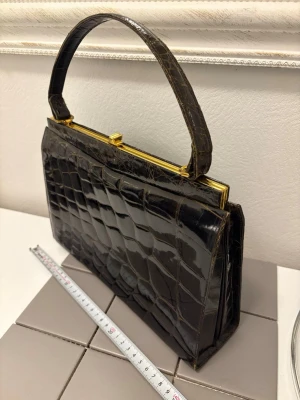 Mörkbrun handväska i krokopräglat skinn - Stilren mörkbrun handväska i krokopräglat skinn med guldfärgad metallram och klassiskt handtag. Väskan har en elegant, rektangulär form och stängs med ett metallspänne upptill. Perfekt accessoar för en trendig look.