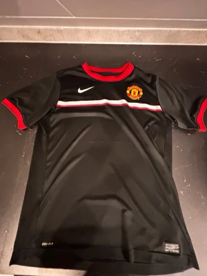 Manchester United tröja - Svart Manchester United tröja från Nike med klubbmärke på bröstet, vit Nike-logga och röd-vit rand över bröstet. Röd krage och röda detaljer vid ärmslut. Tillverkad i Dri-FIT material som andas. Perfekt för fotbollsträning eller support.