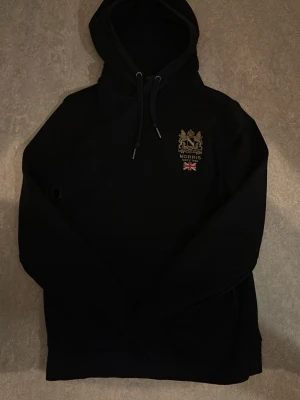 Svart hoodie från Morris - Svart hoodie från Morris med broderad logga och brittisk flagga på bröstet. Klassisk huva med dragsko och känguruficka framtill. Perfekt för en stilren och avslappnad look.