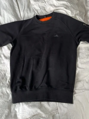 Svart sweatshirt från Alpha Industries - Svart sweatshirt från Alpha Industries med rund halsringning och diskret logotyp på bröstet. Tröjan har ribbade muddar vid ärmslut och nederkant samt en orange insida i nacken. Perfekt för en stilren och avslappnad look.