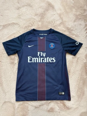 PSG x Nike  - Säljer en blå Paris Saint-Germain matchtröja från Nike med Fly Emirates-tryck på bröstet och klubbmärke. Tröjan har korta ärmar och en röd rand längs mitten. Perfekt för dig som älskar fotboll och vill visa ditt stöd för PSG.