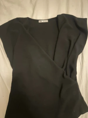 Svart omlottopp från Zara - Snygg svart omlottopp från Zara med v-ringad hals och korta ärmar. Toppen har en stilren design och passar perfekt till många olika looks. Tillverkad i ett mjukt material som känns skönt mot huden.