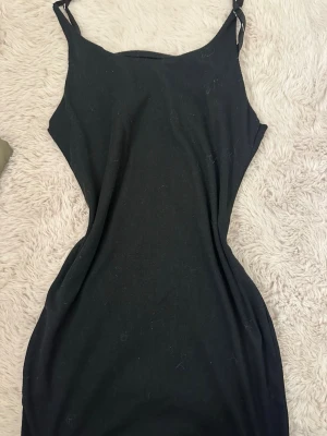 Svart ribbad klänning med smala axelband - En svart, tight ribbad klänning med smala axelband och figurnära passform. Klänningen har enkel design och är gjord i ett stretchigt material som sitter snyggt på kroppen. Perfekt för dig som gillar minimalistisk stil.