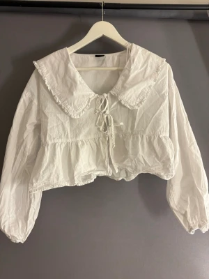 Vit blus med stor krage Gina Tricot - Supersöt vit blus från Gina Tricot med stor volangkrage och knytning framtill. Croppad modell med puffiga långärmar och resår vid ärmslut. Tillverkad i lätt bomullstyg som ger en luftig känsla. Perfekt för en trendig och romantisk look.