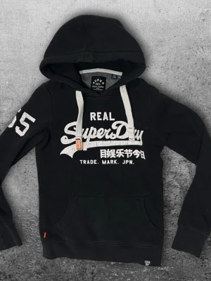 Superdry hoodie XXS - En fet hoodie från Superdry i storlek XXS. Okej skick, ända felet är att trycket inte är i så bra skick. Färg svart! 😀