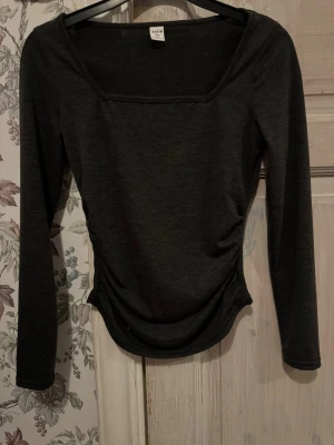 Mörkgrå långärmad topp - En stilren mörkgrå långärmad topp med fyrkantig ringning. Toppen har en figurnära passform och är tillverkad i ett mjukt och stretchigt material, perfekt att matcha med jeans eller kjol.