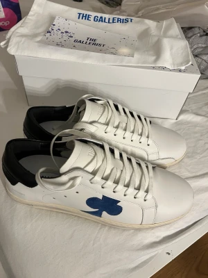 Vita sneakers från The Gallerist - Stilrena vita sneakers från The Gallerist med svart häl och blå klubb-symbol på sidan. Skorna har rund tå, snörning och platt sula. Tillverkade i skinn och levereras med originalkartong och dustbag. Helt nya aldrig andvända. 