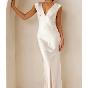 Champagnefärgad långklänning - Elegant champagnefärgad långklänning i satin med djup v-ringning och öppen rygg. Klänningen har en figurnära passform och korta ärmar. Perfekt för festliga tillfällen.