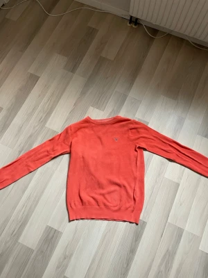 Korallröd stickad tröja från GANT - Stickad tröja i en snygg korallröd färg från GANT. Tröjan har rund halsringning, långa ärmar och ett broderat GANT-logo på bröstet. Materialet är stickat och känns mjukt mot huden. Perfekt för dig som gillar klassisk och stilren design.