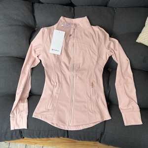 Ljusrosa Define Jacket från Lululemon - Säljer en ljusrosa Define Jacket från Lululemon med slim fit och snygga detaljer. Jackan har dragkedja framtill, två fickor med dragkedja, tumhål i ärmsluten och diskret logga i guld. Materialet är mjukt och stretchigt, perfekt för träning eller chill.Fraktar samma dag också!!