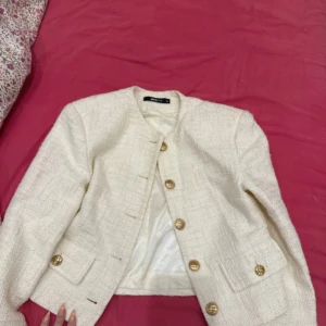 Vit kavajjacka från Gina Tricot - Stilren vit kavajjacka från Gina Tricot i strukturerat tyg. Jackan har rund halsringning, guldiga knappar framtill och på fickorna samt två fickor fram. Croppad modell med lång ärm och helfodrad insida.