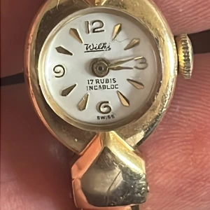 Wilka guldig armbandsklocka vintage för reparation! - Säljer en snygg vintage armbandsklocka från Wilka med guldig metall och vit urtavla. Klockan har markerade siffror vid 12, 3, 6 och 9 samt detaljer i guld. Armbandet är stelt och matchar boetten i guldton. Mekanisk urverk med 17 rubiner och Incabloc-stötskydd.
