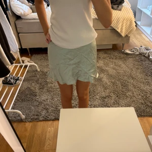 Ljusgrön kortkjol med volangkant - Supersöt ljusgrön kortkjol med volangkant längst ner. Kjolen har en mjuk och glansig yta som ger en fräsch vibe och är perfekt för varma dagar. Passformen är normal och kjolen sitter snyggt i midjan. Enkel att matcha med en basic topp eller sneakers.