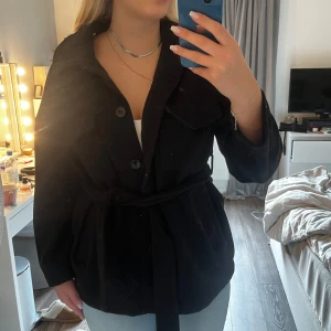 Svart kappa från Gina Tricot - Svart kappa från Gina Tricot med knappar framtill, stora bröstfickor och band i midjan. Original pris:699