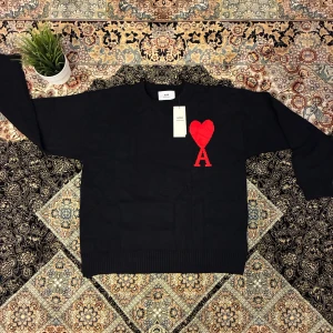  Ami Paris tröja  - Svart stickad tröja från Ami Paris med röd broderad hjärtdetalj och bokstaven A på bröstet. Klassisk rund halsringning och ribbade muddar. Perfekt för dig som vill ha en stilren men ändå unik look.