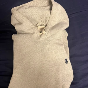 Grå hoodie från Polo Ralph Lauren - Snygg grå hoodie från Polo Ralph Lauren med klassisk broderad logga på bröstet. Tröjan har huva med snörning, magficka och ribbade muddar. Perfekt för chill dagar och enkel att matcha med jeans eller joggers.