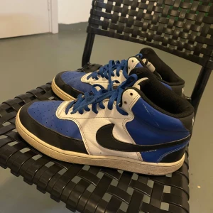 Nike sneakers blå/vit/svart - Säljer ett par klassiska Nike sneakers i blått, vitt och svart med högt skaft och blå snören. Skorna har en rund tå, platt sula och är tillverkade i skinn och syntet. Perfekta för dig som gillar streetstyle och vill sticka ut med en färgstark look. Och vi kan tänka om prisen i chatten!!