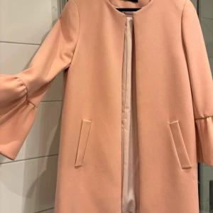  kappa från Zara Woman - Supersnygg  kappa från Zara Woman med raka linjer och volangärmar. Jackan har två fickor framtill och är helfodrad. Perfekt för dig som vill ha en stilren och trendig look i vår eller höst.