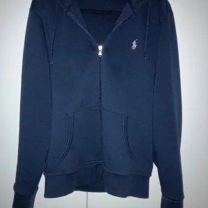 Marinblå hoodie från Polo Ralph Lauren - Snygg marinblå hoodie från Polo Ralph Lauren med dragkedja framtill, huva med snörning och klassisk broderad logga på bröstet. Mjuk bomullsblandning och känguruficka gör den både stilren och bekväm. Perfekt för dig som gillar en clean och fräsch look.