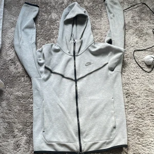 Grå Nike hoodie med dragkedja - Snygg ljusgrå hoodie från Nike med hel dragkedja och svart Nike-logga på bröstet. Tröjan har huva, långa ärmar och svarta detaljer längs dragkedjan och muddar. Perfekt för chill eller träning, tillverkad i mjuk bomullsblandning.
