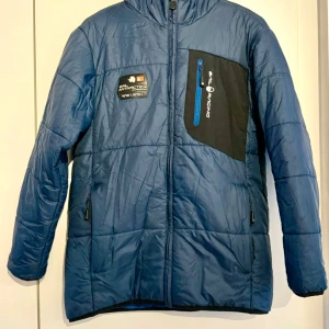 Blå Sail Racing pufferjacka med huva - Blå pufferjacka från Sail Racing med svart bröstficka och blå dragkedja. Jackan har fast huva, två sidofickor med dragkedja och patch med 'Sail Antarctica Expedition' på bröstet. Perfekt för kalla höstdagar och har en sportig vibe. Dunfoder, lättviktsmaterial som andas. Vattentätt yttermaterial som andas. Nypris 1900kr.