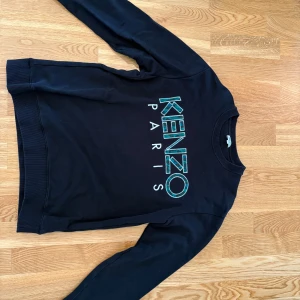 Svart Kenzo Paris sweatshirt - Svart sweatshirt från Kenzo med stort turkost och vitt Kenzo Paris-tryck på bröstet. Klassisk rund halsringning och långa ärmar. Perfekt för dig som gillar streetwear och vill sticka ut med en ikonisk logga.