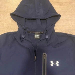 Under Armour vindjacka blå/grå M - Snygg tvåfärgad vindjacka från Under Armour i marinblått och grått. Jackan har huva med snörning, dragkedja framtill och en bröstficka med dragkedja. Under Armour-logga på bröstet och tryck på ärmsluten. Perfekt för blåsiga dagar.
