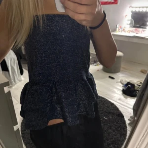 Glittrig mörkblå tubtopp med volang - Säljer en glittrig mörkblå tubtopp med volangkant nertill. Toppen är axelbandslös och har en snygg bandeau-form som sitter tight över bröstet. Perfekt för dig som vill sticka ut med lite extra glitter.