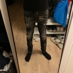 Svarta Dsquared2 jeans med slitningar - Säljer ett par svarta jeans från Dsquared2 med coola slitningar, blekta detaljer och färgstänk. Jeansen har skinny passform och dragkedja i gylfen. Perfekta för dig som gillar streetstyle och vill sticka ut lite extra. På sista bilden så ser ni att knappen är trasig lite men märks inte när de är på.