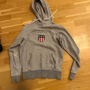 Grå hoodie från GANT med logga XS - Grå hoodie från GANT i mjuk bomull med broderad logga och sköld på bröstet. Klassisk känguruficka, vit snörning i huvan och ribbade muddar. Perfekt för en chill och sportig look. Begagnad skick. Lite slitningar 