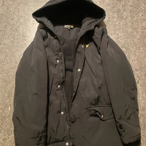 Svart vinterjacka från Lyle & Scott - Svart vinterjacka från Lyle & Scott med huva, dragkedja och knappar framtill. Jackan har två stora fickor med lock och knapp, samt en mindre bröstficka. Klassisk gul logga på bröstet och mjukt foder på insidan. Perfekt för kyliga dagar.
