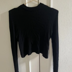 Svart ribbad omlottopp med lång ärm - Säljer en svart ribbad topp med lång ärm och omlottdetalj baktill. Toppen har en hög hals och är croppad i modellen, vilket ger en snygg och modern look. Perfekt att styla med jeans eller kjol för en trendig outfit.